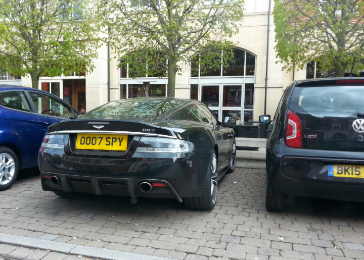 What crappy personalised plates have you seen recently? - Page 428 - General Gassing - PistonHeads - In the image, a parking lot is visible with three cars parked next to each other. On the right side of the image, there are two more cars partially visible. A building with large windows can be seen in the background. The sky is overcast, indicating a cloudy day. The most striking feature is the Aston Martin car on the left with the registration "007 SPY".