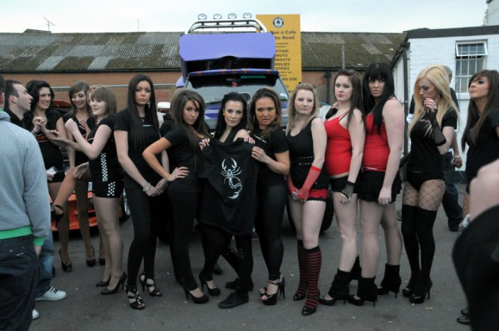 Grid Pistonheads Girlspit Babes - The image shows a group of young women posing near a car with an open trunk. They are dressed in various black outfits, and some have matching hats. The women are holding a spare tire or car tool. The setting appears to be a parking lot, and there are other individuals and vehicles in the background. The image is colorful and seems to capture a moment of camaraderie among the women.