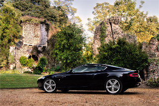 Hello, goodbye & hanging up the pipe and Slippers - Page 1 - Aston Martin - PistonHeads - The image features a small black sports car parked on a gravel driveway in an area with dense vegetation. A green grass field spans the immediate foreground, leading up to trees and shrubberies in the background. A crumbling stone bridge with an arch is visible behind the car, adding a rustic and historical element to the setting. The lighting and shadows suggest it could be during sunrise or sunset.