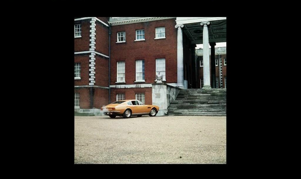 COOL CLASSIC CAR SPOTTERS POST!!! Vol 2 - Page 500 - Classic Cars and Yesterday's Heroes - PistonHeads - The image shows a vintage car parked on a gravel surface in front of two-toned brick buildings, one with red brick and the other with white trim and a darker tone on the lower half. Above the vehicles is a classic architectural structure with Doric columns that resembles a partial portico, suggesting it might be part of a larger building or could be an independent structure on the property. The sky appears to be overcast, and there is a slight blur to the image, giving it a soft focus quality.