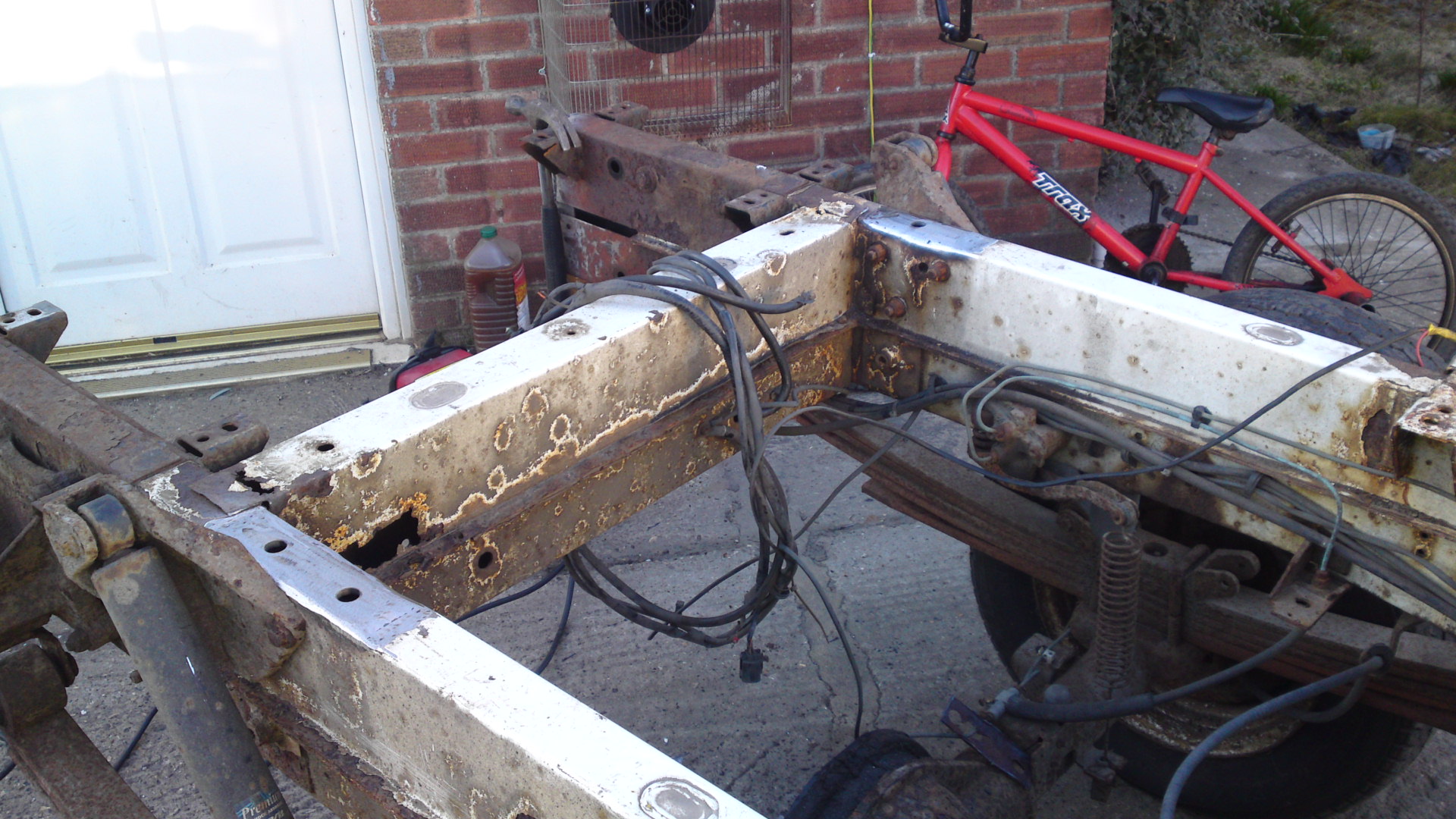 Transporter bed fabrication. - Page 1 - Commercial Break - PistonHeads - The image displays a rusted and seemingly broken metal object, which appears to be the rail of a boiler or stove. It's placed on the floor and has several metal connections and wires attached to it. In the background, there is a red-framed bicycle and a garage door. The overall setting seems to be an outdoor or semi-outdoor garage or workshop, as suggested by the presence of a second bicycle in the corner and other garage-like equipment. There are multiple bolts visible in the object, indicating that it may have been disassembled or is in the process of repair or replacement. The ground shows signs of construction or work, with pieces of material scattered about.