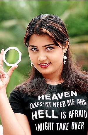 This is a color photograph of a woman posing for the camera. She has dark, wavy hair and is wearing a black t-shirt with humorous text on it. The text reads "HEAVEN DOESNT NEED ME AND HELL IS AFRAID I MIGHT TAKE OVER" in capital letters. She is smiling at the camera and making a peace sign with one hand. The background is blurry and suggests a natural, outdoor setting with greenery.