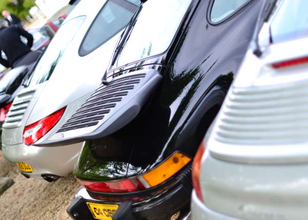 The image showcases a group of cars parked back to back in a parking lot. The cars vary in model and color, indicating a public or shared parking space. They are from different angles, with some cars partially visible in the frame while others are turned around, allowing their rear ends to be seen. The lighting and shadows suggest it's a bright day.