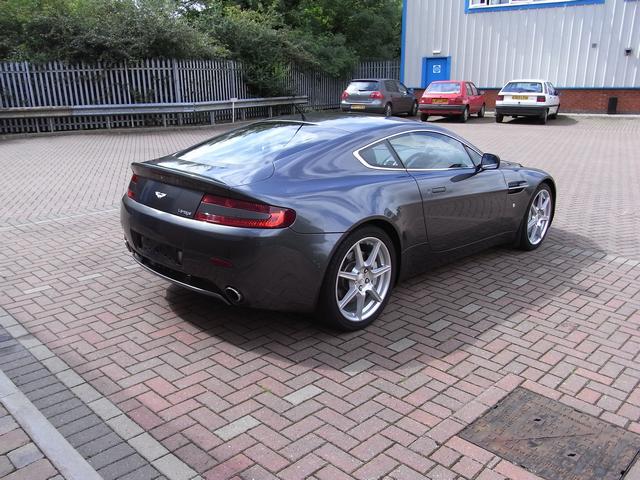 My first Aston - Page 1 - Aston Martin - PistonHeads - The image shows a dark blue sports car parked on a cobblestone surface. The car is positioned at an angle with the front of the vehicle towards the foreground of the image. Its wheels are clearly visible, along with the sleek lines of its aerodynamic design. In the background, two other cars can be seen parked parallel to each other, suggesting the presence of a parking area. Beyond the immediate background, there's an overcast sky and lush greenery, indicating a well-maintained outdoor setting.