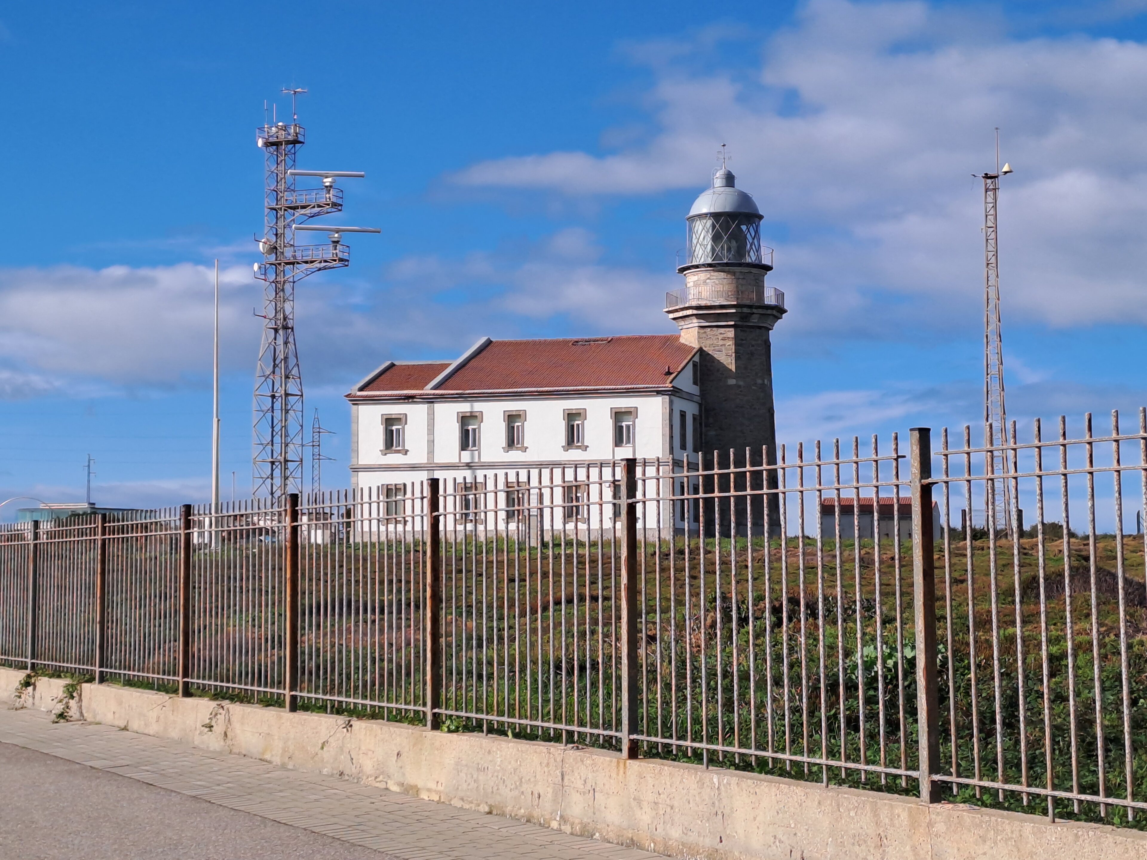 Pistonheads - The image depicts a coastal scene featuring a lighthouse prominently in the center. The lighthouse is an old-style structure with two visible levels, topped by a cupola and a tall mast or antenna. It's painted white, contrasting with its dark roof and windows, and stands out against the sky.

The lighthouse is situated within a gated area that appears to be in a state of disrepair; the fence surrounding it seems weathered and old, contributing to a slightly dilapidated atmosphere around the structure.

In the foreground, there's a small fence running parallel to the road, suggesting that the road is quite close to the lighthouse. A person can be seen walking by on the right side of the image. The sky is clear and blue, indicating a sunny day.

The overall scene suggests a coastal town or village, with the lighthouse serving as a landmark for navigation purposes.