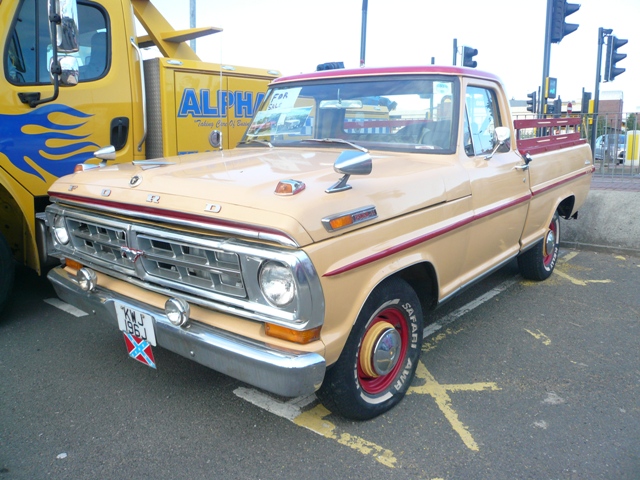 Metrocentre Pistonheads - The image showcases a beige Ford Bronco parked by the side of the road. The truck is colored a light beige with distinct maroon and black accents on the front and sides. The Bronco is equipped with numerous identification markings, including a license plate that reads "KJW 567". There is a red arrow pointing towards the truck with the inscription "ALPHA". The truck is parked neatly near the curb, and the road is devoid of any other traffic, giving the impression of a quiet street.