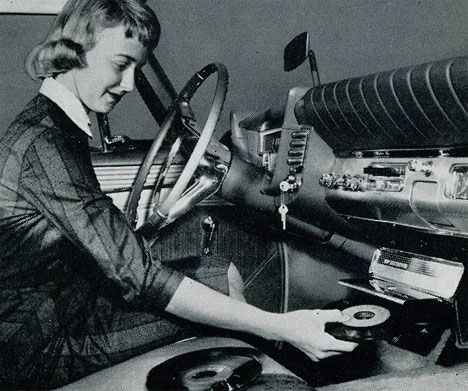 What was the most ridiculous car accessory in history? - Page 4 - General Gassing - PistonHeads - The image is a black and white photograph of a vintage car, featuring a woman seated in the driver's seat. The car's interior contains classic car accessories such as a traditional radio and key ignition. The woman appears to be in a relaxed posture and is looking directly at the camera. The rearview mirror, a prominent feature in vintage cars, is situated in front of her. On the right side of the photograph, a jukebox is visible inside the car, adding to the nostalgic feel of the scene.