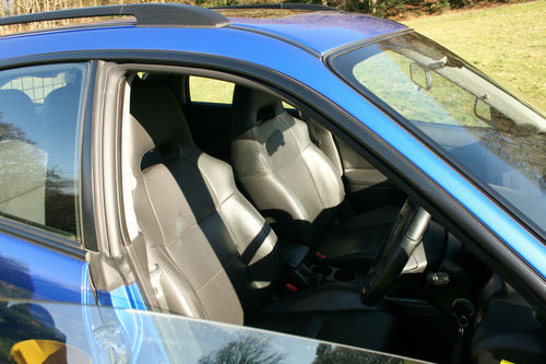 A car is parked on the side of the road - Pistonheads - The image showcases the interior of a compact car. The car has a striking blue exterior and a contrasting black interior. The vehicle features two black seats visible from the front door. The steering wheel, matching the seats in color, is positioned centrally. The passenger side door is open, revealing a clear sky outside.