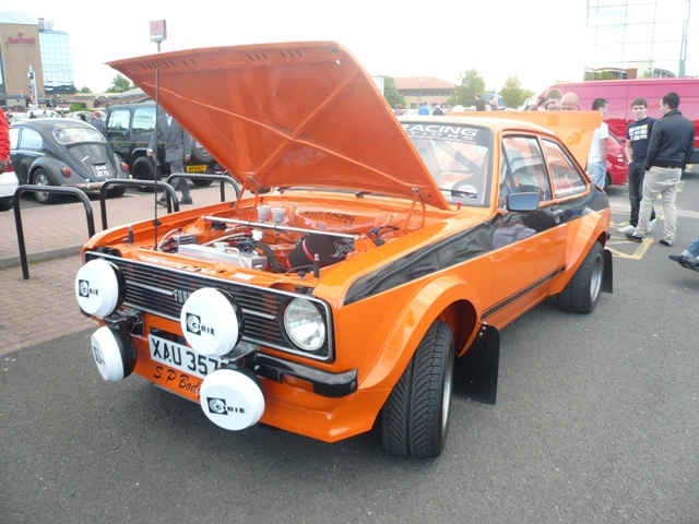 Pistonheads Metrocentre - The image features an orange modified car with its hood open, parked in a parking lot. A light-colored rally car is parked next to it. There's a group of people observing the cars, with a couple of young individuals in particular leaning into the scene. Some of the surrounding vehicles also appear to be vintage or modified, indicating that this might be a car show or meetup.