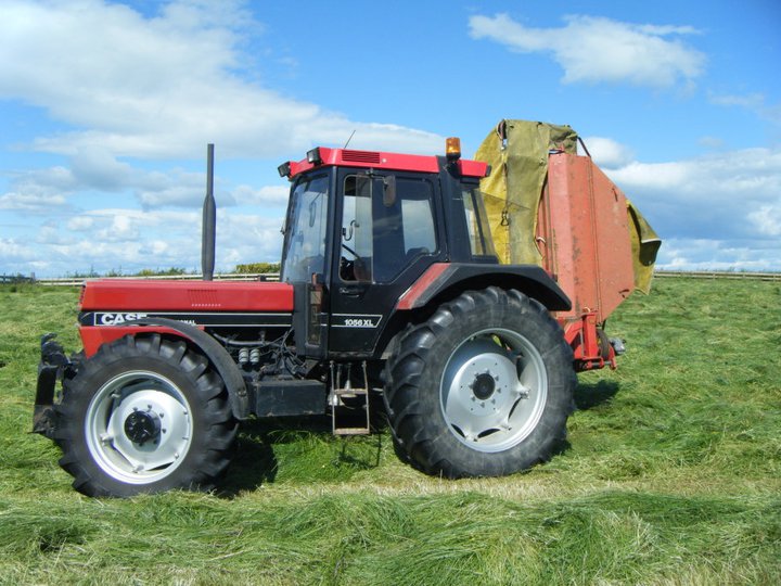 Classic tractors - Page 3 - Classic Cars and Yesterday's Heroes - PistonHeads - In the image, we see a red and black Case IH farm tractor parked in a field covered with lush green grass. The tractor, which is displayed prominently in the foreground, has its front loader attachment attachment hooked up, indicating that it might be ready for agricultural work. The sky above is mostly clear, suggesting it might be a sunny day. In the background, there is a trail-style bridge, which can also be described as a train overpass, indicating that the field is close to or part of a train route. The overall environment suggests a peaceful and productive countryside setting.