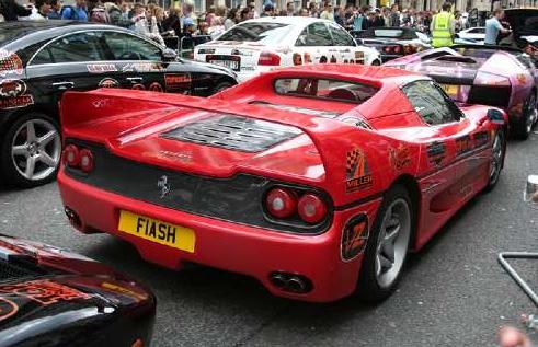 - Page 530 - General Gassing - PistonHeads - The image captures a vibrant scene on a street where several brightly colored sports cars are parked. The most striking is a red Ferrari, which stands out against the others. Various other cars can be seen around it, some of which appear to be race cars. In the foreground, a licence plate on one of the cars reads "FIASH". The setting seems to be a populated area with people visible in the background. Some cars in the background have advertisements on them, adding to the bustling, energetic atmosphere.