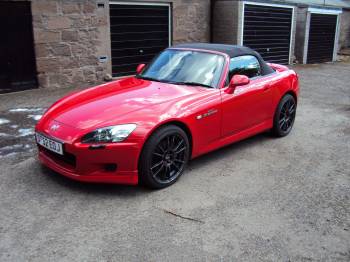 Just bought an S2000 :) - Page 1 - Honda - PistonHeads - The image shows a sports car parked on a gravel surface. The car is red with black accents and appears to be a convertible, based on its design and the retractable hard top roof. The setting seems to be in a driveway in front of a garage, suggesting that this could be the owner's home or a dedicated area for parking vehicles. There are no people in sight, giving the scene a still and deserted feel. The overall picture presents a solitary scene of a luxury car parked against the backdrop of a garage, exuding a sense of opulence and quietude.