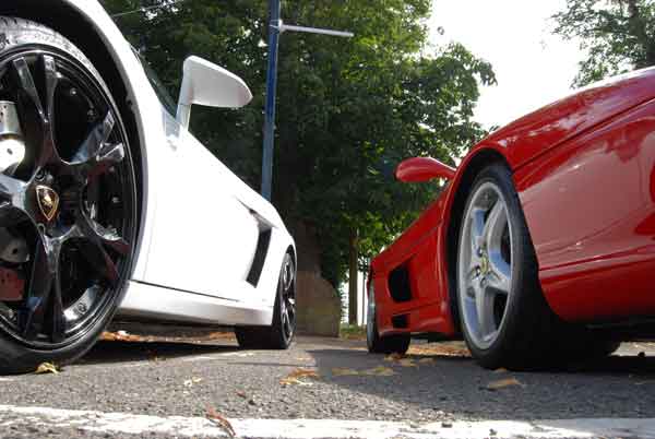 A motorcycle parked in front of a building - Pistonheads - The image presents two luxury sports cars parked side by side. The cars contrast in color: one is white, and the other is a deep, glossy red. The white car, positioned on the left, has a large, prominent rear wing, adding to its aerodynamic appearance. On the right, the red car mirrors the white one's sleek design, with a smaller rear wing. The backdrop of the scene is a serene setting of lush greenery. The cars appear to be parked on a road or a driveway, showing that they are ready for a ride at any moment.