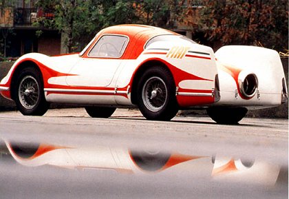 Pistonheads Quizanyone - The image features a vintage racing car on a wet roadway. The car is predominantly red and white, with a singular large wheel visible in the foreground. In the background, another similar car is partially visible in profile. Trees and a partly visible building flank the scene, suggesting a semi-urban or park-like setting. The wet roadway reflects the car and the surrounding environment, adding a vibrant reflection to the scene.