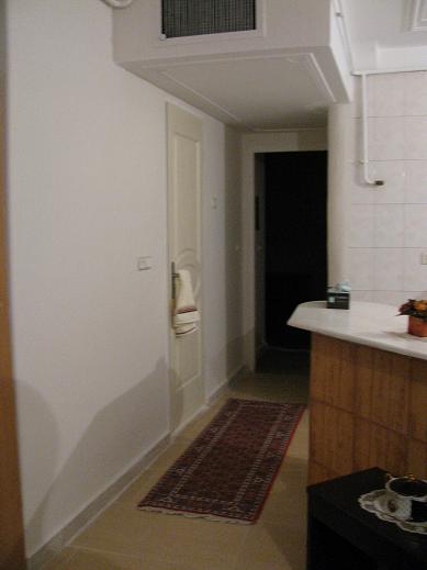 The image portrays a kitchen that is typically separated from the dining area by a countertop. The kitchen is neat and tidy with a white oven on one side and a wooden cabinet on the other, indicating a well-maintained and clean space. A rug is spread out on the floor in front of the oven, possibly for comfort or cleanliness when cooking. A vase is visible on the countertop, and there are pots, further suggesting this as a fully functional kitchen. The lighting is dim, and the photo is taken from the viewpoint of the kitchen entrance.