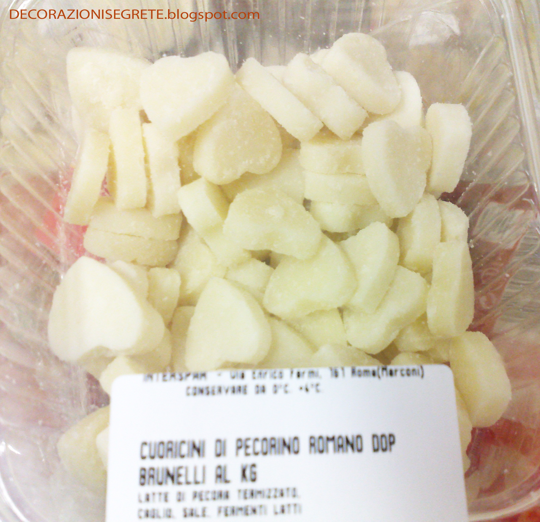 The image shows a clear plastic container filled with diced, white, pear-shaped fruits, which appear to be dessert pears or possibly chunks of white fruit like quince or melon. On top of the fruit, there is a white label with black text that contains the product name "CUORICIN DI PECORINO ROMANO DOP." The label also specifies the quantity as "1/2 KG." Below the label, additional text suggests that this is an organic or certified fruit product. In the background, the container is placed against a wooden surface, and there is a website address "decorazionisi molto belle.blogspot.com" partially visible at the top of the image. The overall setting seems to be in a kitchen or home environment.