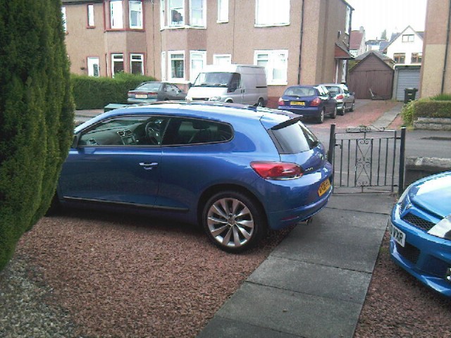 Pistonheads Vxr Jealous - This image shows a close-up view of the side of a blue car parked on a stone driveway. The car is a hatchback model with a sunroof and alloy wheels. Behind the car, there is a black wrought iron gate, and in the background, a residential street is visible with several parked cars and houses. The photo is taken during daylight with clear visibility.