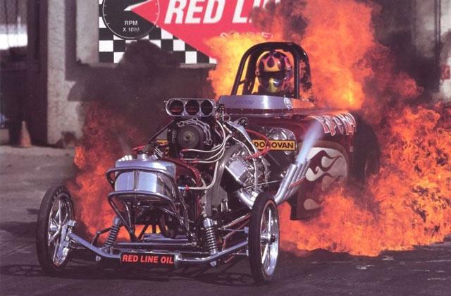 Fantastic Motorsport Photos! - Page 121 - General Motorsport - PistonHeads - The image showcases a thrilling scene of a car participating in a drag race. The car, emblazoned with the text "Red Line Oil" and other sponsor logos, is in flames, indicating high performance or an engine malfunction. The fire partakes in all three dimensions, engulfing the rear of the car and rising up into the air. In the background, a race track sign labeled "DRAG" is visible, reinforcing the context of the event. The car's driver can be seen through the open cockpit, seemingly in the midst of the action.