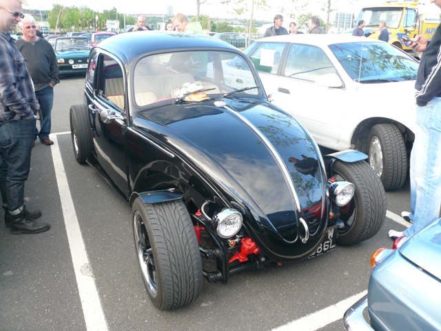 Metrocentre Pistonheads - This image captures an automotive gathering where a black Volkswagen Beetle car is in the foreground with other cars parked in the background. The VW Beetle, a two-door convertible, stands out due to its iconic shape, fastback roof, and red engine exposed through a scoop on the hood. A person is seen in the background looking at the car, indicating interest and admiration for this classic vehicle.