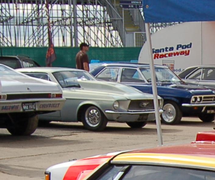 Pistonheads Silverstone Classic Goto - The image depicts an outdoor scene at what appears to be a racing event or a car show. A variety of vintage cars are parked in a lot, with street signs and barriers visible behind them. People and spectators are present amidst the parked cars, engaging in the activity. One individual is walking near the cars, and two others are standing closer to the camera. There's a tent structure in the background, adding to the setup of the event.