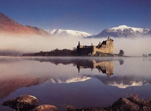 Summer Pistonheads Tour - The image presents a serene landscape dominated by the natural beauty of a mountain-filled skyline and a body of water that mirrors the sky and the distant castle. The foggy atmosphere adds a mystical quality to the scene, highlighting the silhouette of the castle and the ruggedness of the mountains. The castle, reflecting majestically in the calm waters, and the towering mountains stand as timeless landmarks, exuding an air of antiquity and grandeur. This peaceful scene, bathed in the soft light of what appears to be either early morning or late afternoon, evokes a sense of tranquility and awe at the natural and architectural wonders depicted.