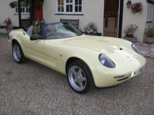 Caterham Pistonheads - The image features a yellow Alfa Romeo sports car parked at the curb in front of a house. The vehicle's sleek lines, unpainted trim, and black rubber tires are prominently visible. The convertible top is down, providing a clear view of the interior. The surroundings include potted plants adding a touch of greenery to the scene.