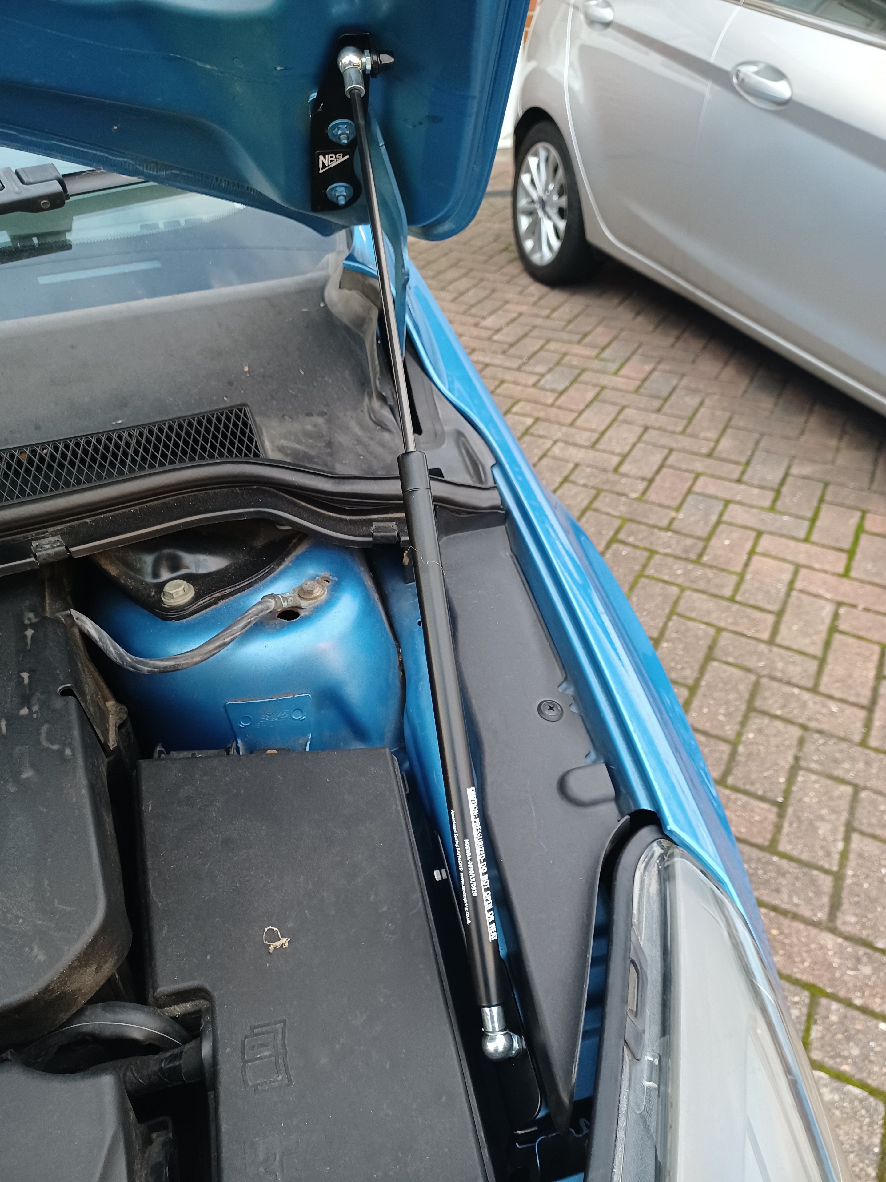 The image shows a blue car with its trunk open. Inside the trunk, there is a collection of items including a suitcase and what appears to be a toolbox or storage compartment. There are also various tools and components visible, suggesting that some maintenance work may be taking place. A gas can is located on top of one of these components. The car is parked on a brick driveway, and in the background, another vehicle can be seen.