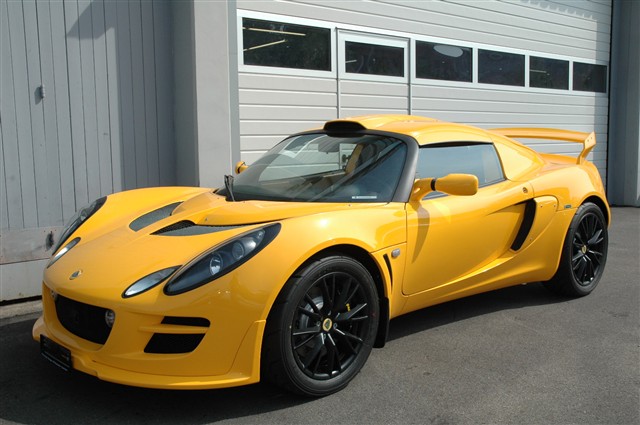 Pistonheads - A bright yellow sports car parked in front of a building. It is daytime. The car has sleek curves and sports car styling. The paint is shiny, which reflects the sunlight. The background features a gray building with a series of windows and a closed garage door.