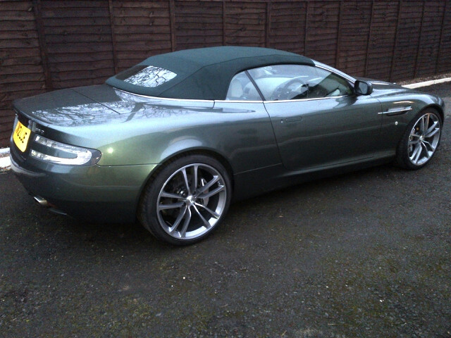 DBS wheels on DB9 - (more or less) - Page 1 - Aston Martin - PistonHeads - This image features a sleek, metallic gray sports car parked on a paved driveway. The car's body gleams, highlighting its aerodynamic design. The soft-top roof is down, revealing the interior as empty. The vehicle's front wheel is visible, showcasing the car's design and the reflections of the surroundings. In the background, a wooden fence runs horizontally across the frame. The car's style suggests it's a convertible, particularly given the open roof.