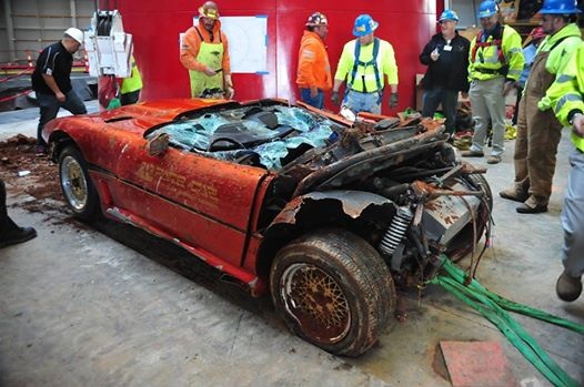 1,000,000th. Corvette returns from chasm - Page 1 - Corvettes - PistonHeads - This image captures a scene of a severe car accident in a construction or industrial setting. A red, rusted car is severely damaged, with parts of the vehicle ripped off and scattered around the area, and its doors are shattered. A group of people, including workers or bystanders, are observing the aftermath of the accident. They are dressed in various types of work attire, with some wearing hi-vis vests and others in regular clothing. The overall atmosphere and condition of the car suggest a recent and violent collision, likely involving considerable force.