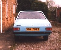 Popular Escort Pistonheads - The image shows a blue-green car parked on a gravel area with a blurred brick house in the background. The vehicle features yellow and black license plates. The photo has a vintage or nostalgic feel due to its grainy texture and the attire and hairstyles of the person partially visible on the left side of the image, suggesting it might have been taken in the past.