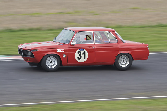 Photography at Castle Coombe - Page 1 - South West - PistonHeads - The image captures a vintage racing car in action on a racetrack. The car is painted red and has a distinctive number '31' prominently displayed on its sides. It's clearly designed for high-speed performance, with sleek aerodynamics that suggest it's a track-only vehicle. The driver's hands are visible, gripping the steering wheel as they navigate the track, which curves subtly ahead. The car's advanced design and the intensity of the driver's focus hint at a moment of high speed and competition.