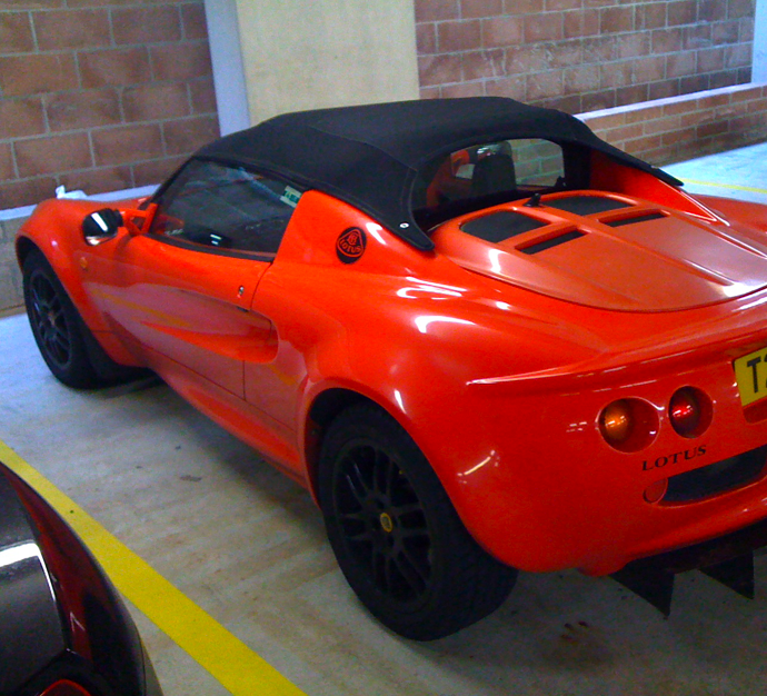 Decal width and colour - Page 1 - Elise/Exige/Europa/340R - PistonHeads - The image features a vibrant orange Lotus sports car parked in the middle of a parking lot. The car is characterized by a sleek, convertible design with a black soft top that's up. It has a small flat trunk at the rear and features a visible logo on the side. The setting appears to be an indoor parking area, with other cars visible behind the Lotus, suggesting a multi-story parking structure. The sunlight streaming in suggests a bright, possibly sunny day outside.