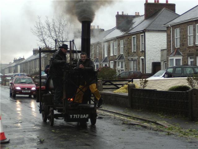 Oldest design here ? - Page 1 - Classic Cars and Yesterday's Heroes - PistonHeads - In this image, two individuals are enjoying an old-fashioned, steam-powered locomotive ride on a street. The locomotive is the main focus of the image, with a plume of steam pouring out of its chimney. It's being pulled by a cart with two wheels, which is carrying the passengers. The street they're on appears to be in a residential area, as evidenced by the houses and parked cars visible in the background. The overall scene suggests a fascinating blend of the past and the present.