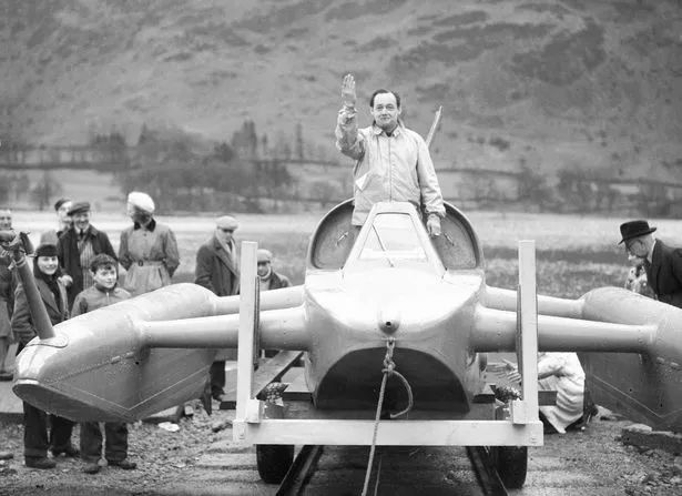 Bluebird K7 Latest - Page 4 - Boats, Planes & Trains - PistonHeads UK - This black and white photograph captures an outdoor scene featuring a man standing atop a peculiar, futuristic-looking vehicle. The man, wearing a white shirt and tie, is raising his left hand in a wave or greeting gesture. Surrounding the man are several onlookers, including adults and children, who seem to be engaging with the vehicle or the event that is taking place. The background consists of rolling hills and a misty landscape, suggesting a rural or nature setting.