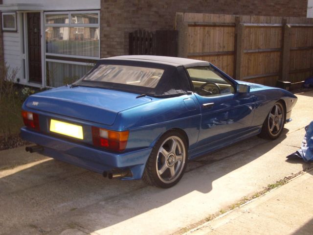 A car parked on the side of the road - Pistonheads - The image depicts a vintage blue sports car parked on a driveway. The car is positioned on a rectangle of concrete, with the large headlights prominently visible, and the windows showing reflections of the surroundings. Behind the car, there is a wooden fence and a semi-closed garage door, providing privacy and security. The setting appears to be a quiet residential area, possibly on a sunny day given the clear visibility and soft shadows. The car's design is suggestive of an era when sports cars were predominantly rear-wheel drive with a hardtop or convertible roof, commonly powered by high-revving, fuel-injected engines.