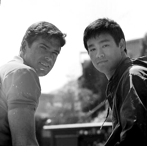 This black and white image captures a candid moment between two young men. They are standing close to each other, engaging in an animated conversation. The man in the foreground is dressed in a casual shirt and is looking at the camera with a slight smile, whereas the other man in the background is wearing a jacket and is more focused on the background, suggesting he might be pointing at it. The background itself is washed out and nondescript, putting the emphasis on the subjects and their interaction.