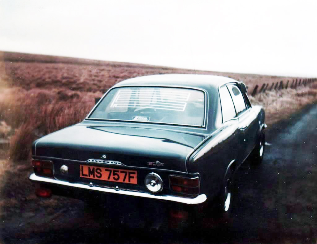 Pistonheads - The image is a photograph of an old-model, two-door car parked on what appears to be a rural road. It's a black and white shot that gives the scene a timeless quality. The car has a prominent front grille and round headlights, characteristic of its era. There's no visible text or branding in this image. The road seems to be in a countryside setting with sparse vegetation on either side, suggesting a remote location.