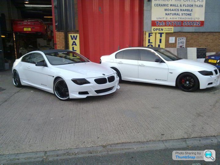 Badly modified cars thread - Page 134 - General Gassing - PistonHeads - The image shows two BMW coupe cars parked side by side in front of a red building. Both cars are white with darker color accents, and they are positioned close to each other, taking up the majority of the wide-angle shot. The tire design on both cars is similar, with a black and silver color scheme. Behind the cars, the bay has a "WELL" sign, and there are additional signs suggesting that this is a workshop, as well as a sneaker store, and some service equipment visible. The ground is paved, and the picture appears to be taken in the daytime.