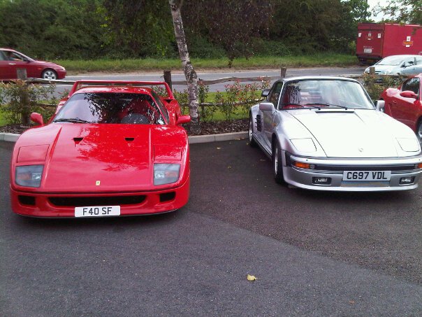 The Hare, 6th August - Page 1 - Kent & Essex - PistonHeads - The image features a parking lot with two vintage sports cars. The red Ferrari is on the left side of the frame, with a prominent nose, round headlights, and a black front grille. It wears a license plate that reads "FA40 SF." The silver, possibly Audi, sports car is on the right, distinguishing itself with round headlights and a rectangular grille design. The license plate on this car is "C69VLD." The environment suggests a rural setting with a fence and greenery in the background.