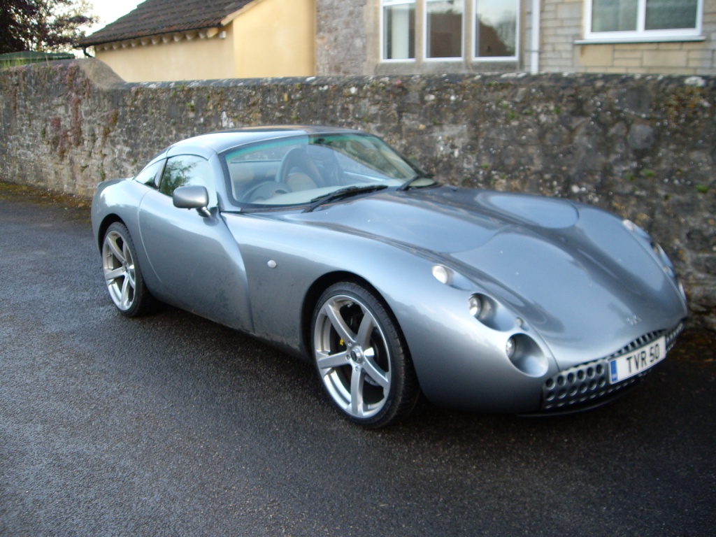 Pistonheads Tunnel Bristol - The image features a sleek gray sports car parked on a wet pavement. The car is positioned at an angle and takes up most of the frame. The car's design includes a striking aerodynamic body, which adds to its futuristic and sophisticated appearance. The surrounding environment consists of a stone wall in the background and a house to the left, both of which provide a contrasting backdrop to the shiny car. The wet road reflects the car and the stone wall, giving the scene a glossy and dynamic quality. The overall image presents a tranquil setting with the sports car being the focal point.