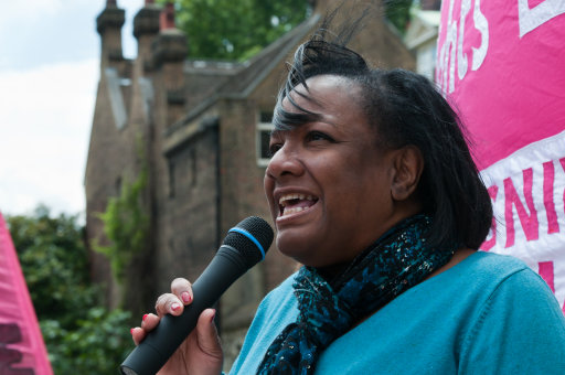 Jeremy Corbyn Vol. 2 - Page 511 - News, Politics & Economics - PistonHeads - The image is a color photograph featuring a woman in the foreground who appears to be speaking into a microphone. She is wearing a bright blue top and has her mouth open, possibly in the midst of speaking. Behind her, there's an outdoor setting with buildings and greenery visible in the background. The woman has her hair tied back and is looking up and to the right, suggesting she is addressing an audience or engaging with someone or something outside of the frame. There's a sign or banner visible in the background with text, but the resolution and angle make it difficult to read the full message.