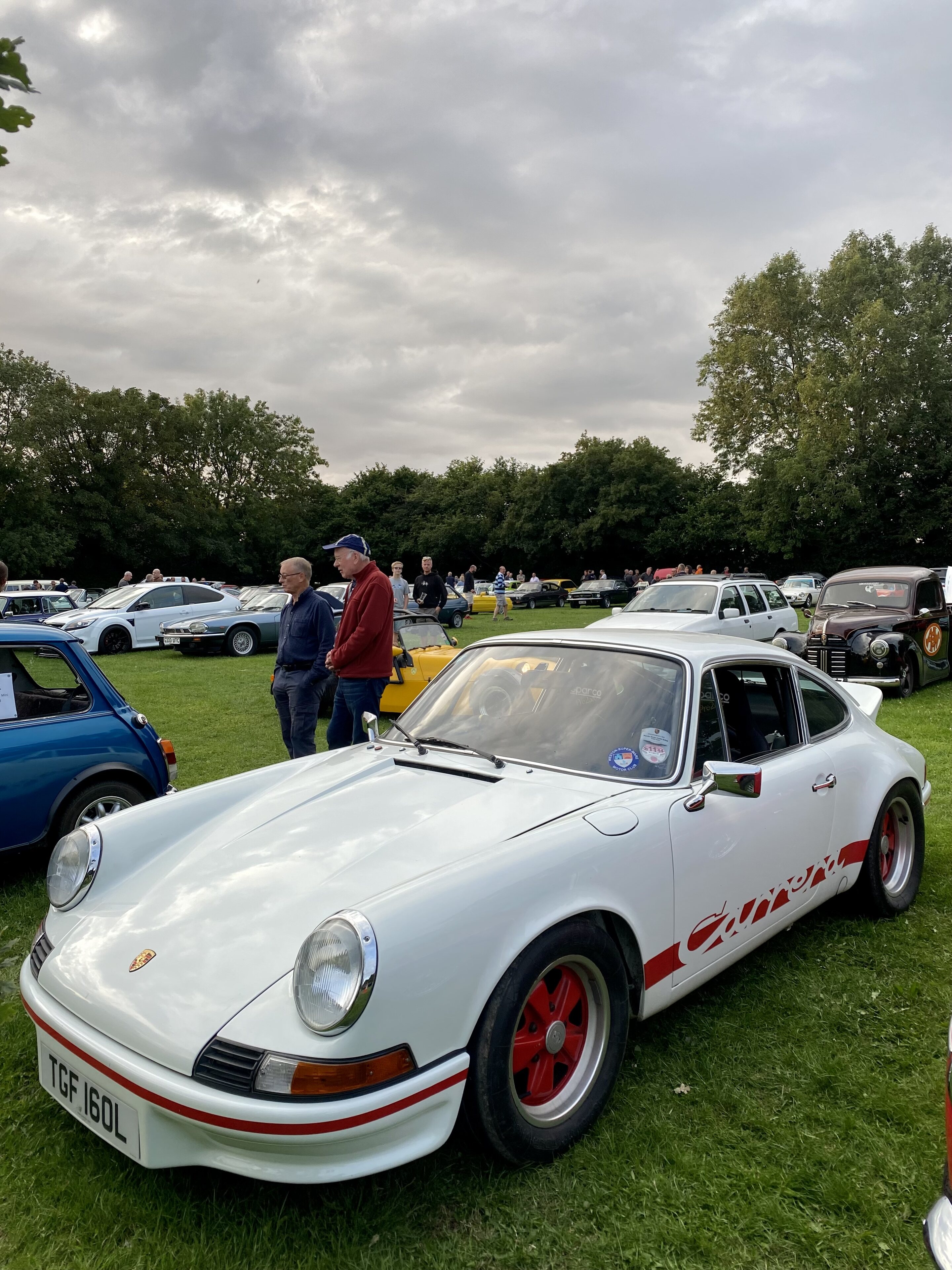Pistonheads - The image is a photograph of an outdoor scene where vintage cars are parked on grass. There is a white Porsche with red accents and a black roof, positioned to the right of the frame. Adjacent to it, there is another classic vehicle with a yellow paint job and a matching yellow roof. In the background, several other cars can be seen, including one with a blue top and a cream-colored car with a white roof and stripes on its side. There are also people present in the background. The sky overhead appears to be overcast with some patches of sunlight. The setting suggests this could be an auto show or a gathering for classic car enthusiasts.