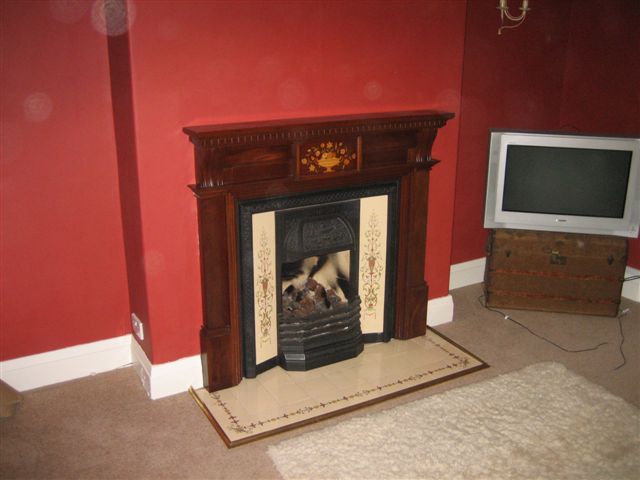 Installing a wood burner, DIY or Pro?  - Page 2 - Homes, Gardens and DIY - PistonHeads - The image depicts a living or common area characterized by a cozy atmosphere. A fireplace, featuring a decorative mantel and a carved right-hand side, is the focal point of the room with its black and white tiles as well as a chevron pattern around the perimeter of the hearth. The television is positioned to the right of the fireplace, atop a piece of furniture that appears to have drawers. To the left of the fireplace, part of a cowhide rug is visible, adding to the rustic design aesthetic of the room. The walls of the room are painted red, which complements the rug and the overall ambiance of the space.