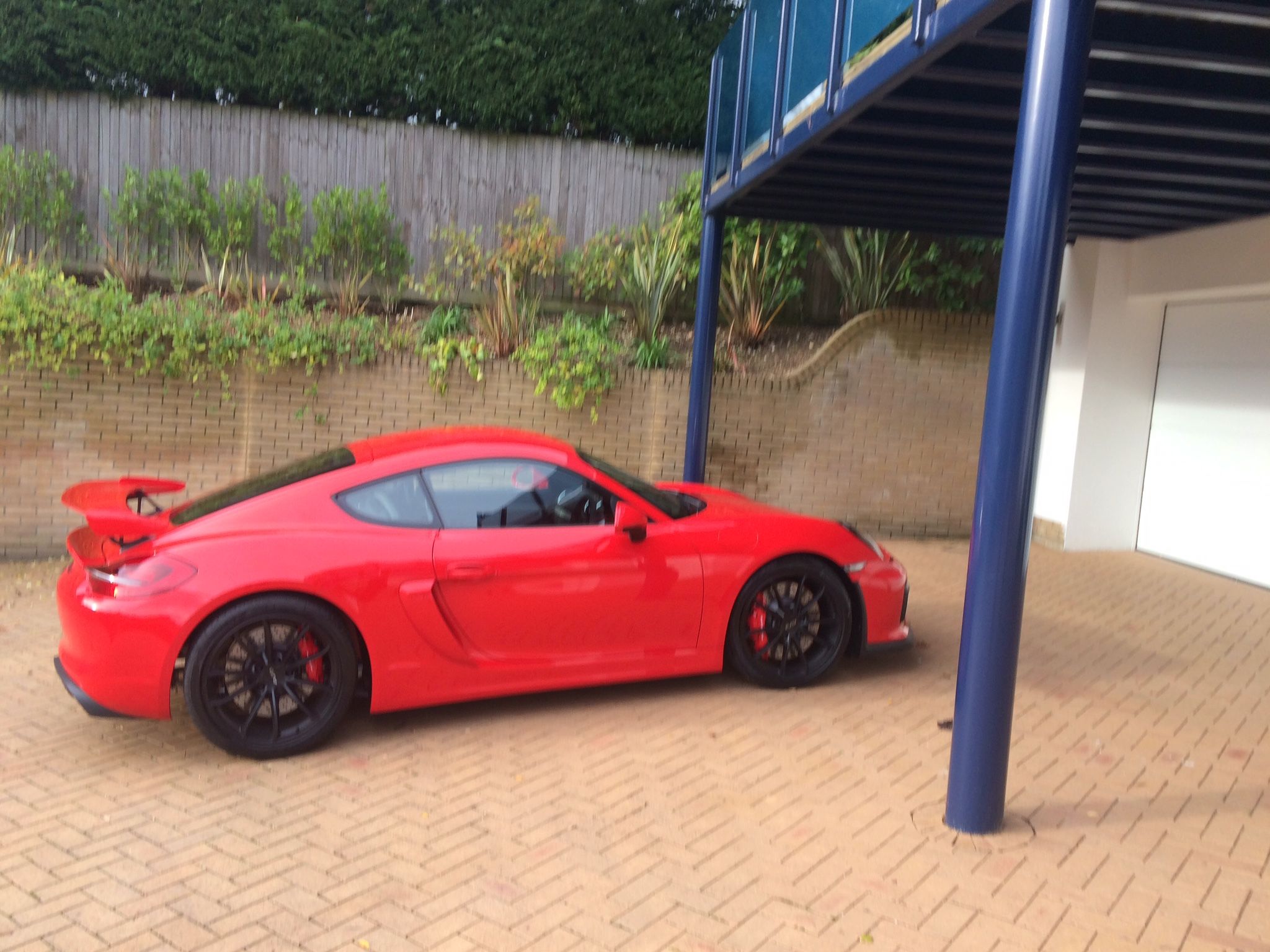 Prospective 981 GT4 Owners Discussion Forum. - Page 443 - Porsche General - PistonHeads - The image shows a red Porsche sports car parked on a brick paved driveway. The car is positioned under a blue balcony structure, indicating it is likely a residential setting. The vehicle's rims are a contrasting black, matching the hood scoops and side mirrors. The setting suggests a quiet, suburban environment with greenery visible in the background. The brick driveway has a neat appearance, adding to the overall tidiness of the scene.