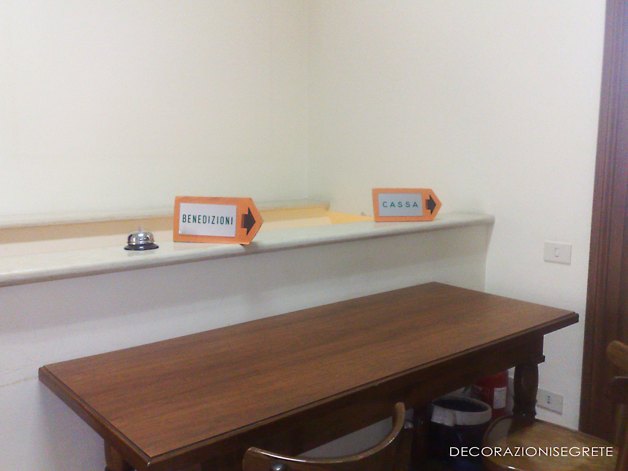 The image shows an interior space with a desk placed against a wall. On the desk, there are two clear plastic sign holders attached to its surface. The holders each feature a small plate bearing a printed declaration saying "ben prego di?enedizione" in bold, black letters. The room appears to be well-lit and is furnished with minimal decorations, suggesting a functional or utilitarian setting. The overall style of the photograph is candid and straightforward, capturing the details of the desk and signs in a clear, unembellished manner.