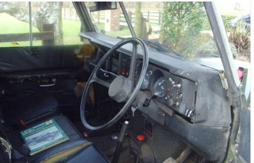 Pistonheads Repurchased Landy Fathers Day Present - The image captures the interior of a vintage vehicle, specifically the driver's seat area. The vehicle appears to be in need of restoration, as indicated by its old and possibly damaged appearance. A steering wheel, worn out from use, is prominently positioned in the center of the image. To the right, there's a manual transmission shifter, and a book with the cover title "POW How to Survive" is resting on the seat, further enhancing the historical and possibly wartime context suggested by the vehicle's style. A window with the screen raised separates the passenger area from the outside.