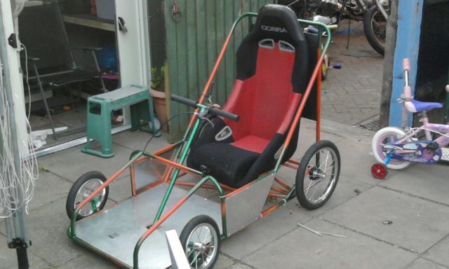 A bike parked next to a wooden bench - Pistonheads - The image features a unique, homemade go-cart. The go-cart is made from a utility trailer or a lawn mower cart, with a red seat and black seat belts attached in the center. It stands on four wheels, two at the back and two at the front. The go-cart appears to be in a somewhat cluttered or organized garage. The trailer is partly yellow with green highlights, suggesting it might be an old lawn mower with a poly cart attachment. A red and blue streamer is attached to one end of the trailer, possibly as a decoration or to enhance visibility. A bicycle is parked next to it on the right side. The trailer is positioned in front of a large green door. Several other vehicles, including motorcycles and bicycles, are scattered in the background.