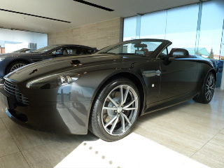 Owner Vantage Pistonheads - The image depicts an interior showroom, with the central focus being a luxurious, dark-colored sports car. The car is positioned for an optimal viewing angle, showing off its sleek lines, distinct design features, and alloy wheels. The showroom itself has a spacious, modern feel, with a large window that allows light to flood into the space and vehicles on display behind the sports car. The lighting accentuates the car's color and the polished floor reflects it, creating a refined and stylish atmosphere.