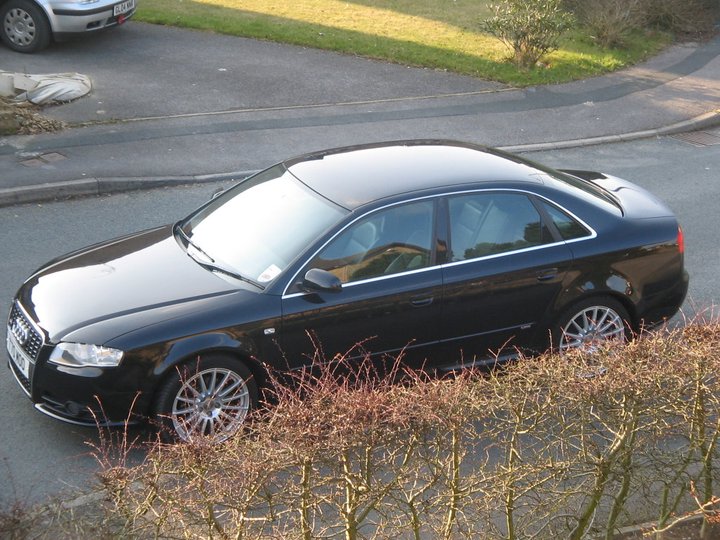 RE: MG ZT-T V8 260: Spotted - Page 7 - General Gassing - PistonHeads - The image shows a dark-colored Audi sedan driving down a street at a slight angle to the camera. The car appears to be in motion, with sunlight glinting off its body and through its side windows. The photo is taken from an elevated perspective, looking across a lawn or garden to the parked car. The car is positioned parallel to the curb of the street, and there is some reflection of the car on the shiny surface of the street.